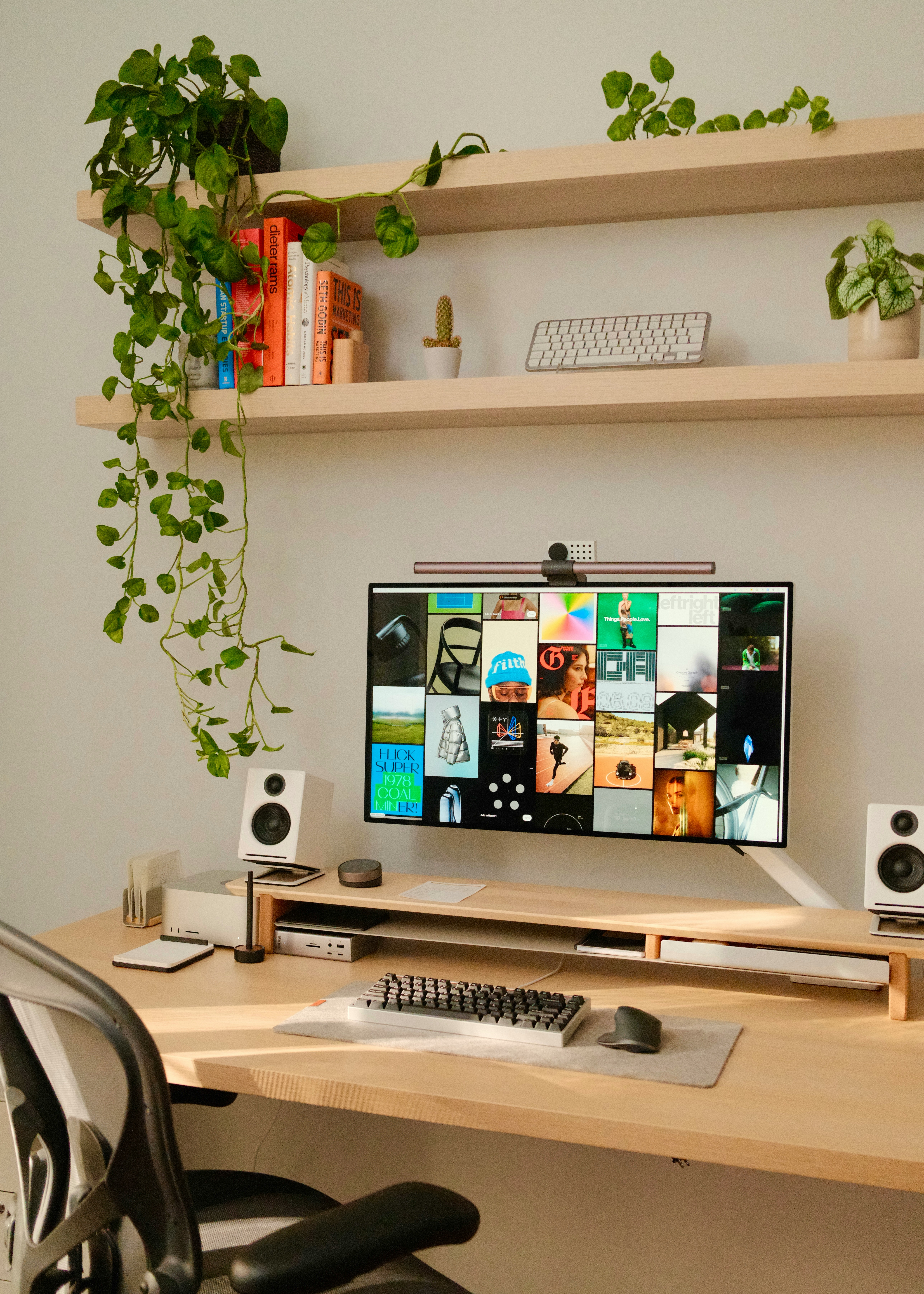 Studio workspace with a monitor showing a portfolio grid, shelves with plants and books, and speakers on a light wood desk